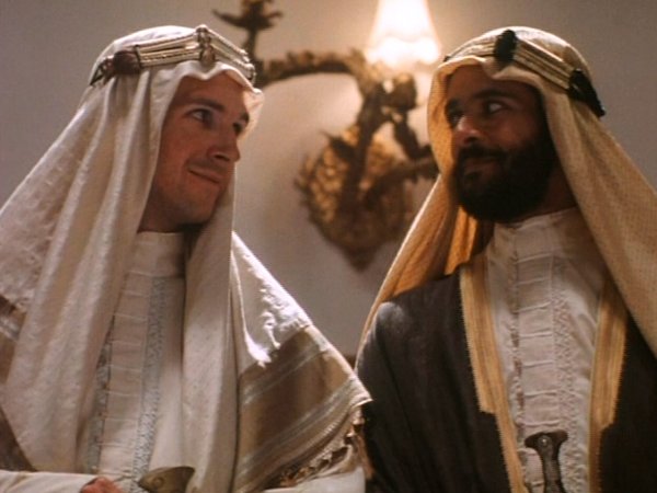 Ralph Fiennes, a white man with a barely-there mustache, and Alexander Siddig, a man with tan skin and a full-but-short black beard and mustache, look at each other warmly. Ralph is wearing a white keffiyeh and matching white robes. Siddig wears a light brown keffiyeh and robes that are dark and light brown and white.