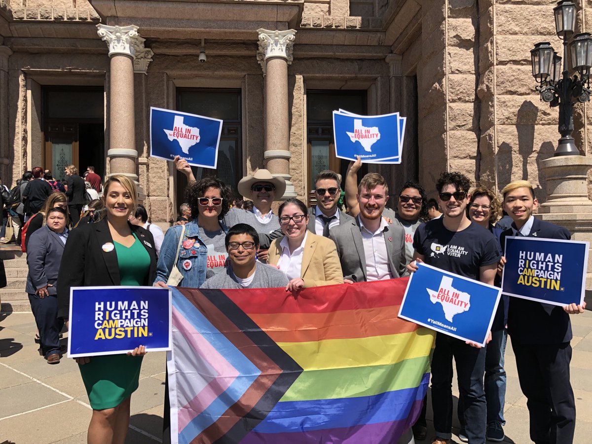 11 people pose behind a rainbow flag (with the ROYGBV stripes horizontal and a triangle made out of black, brown, light blue, pink and white colors). Some are holding HRC signs. All are smiling. One is wearing a cowboy hat and proudly holding two signs!