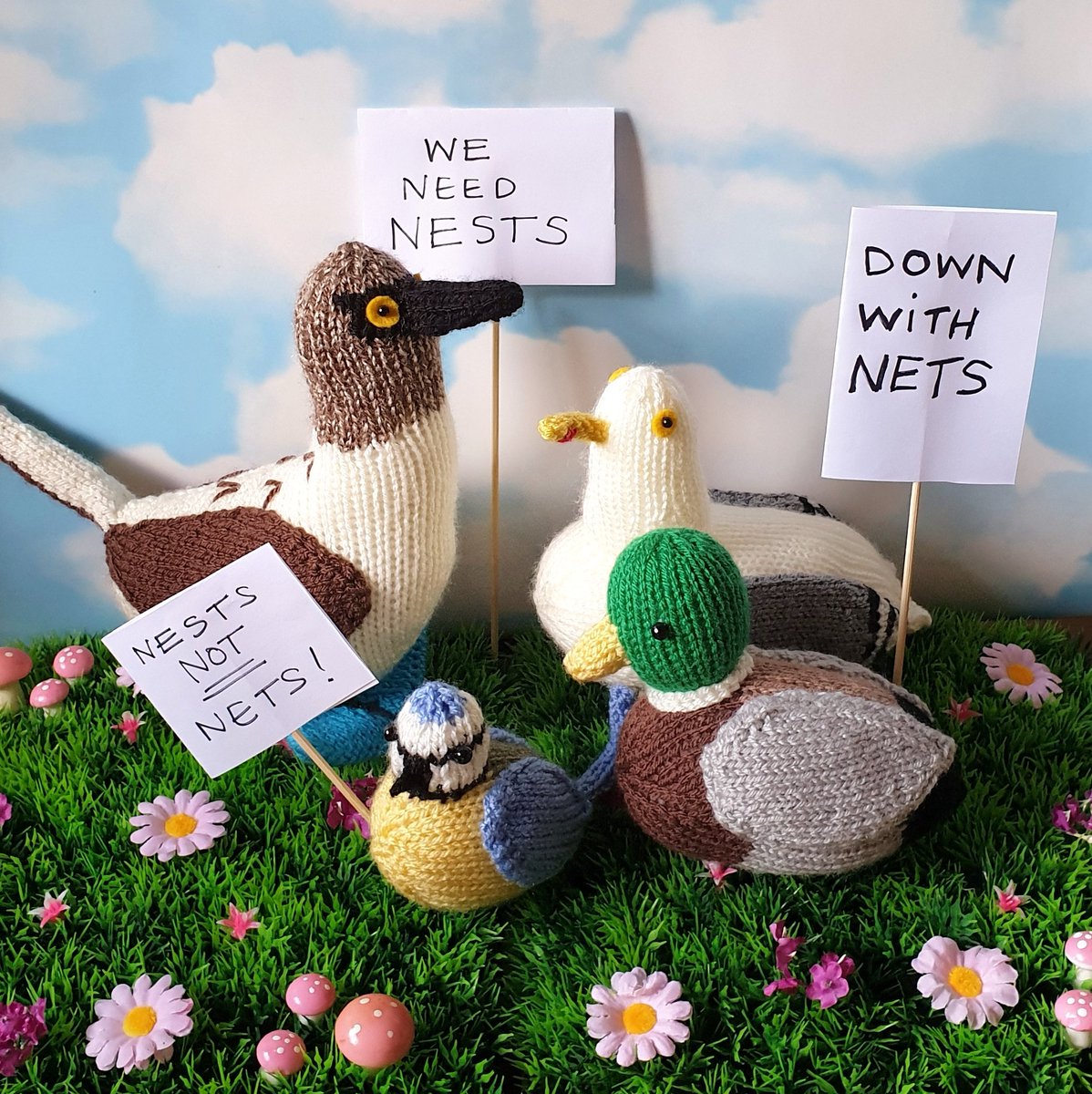 a group of knitted birds protesting with signs saying we need nests, down with nests and nests not nets
