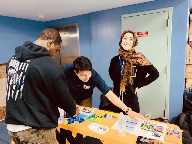 The image shows a man in a sweatshirt standing next to a table with a golden tablecloth, a man reaching to pick up a flyer, and a woman smiling on their right.