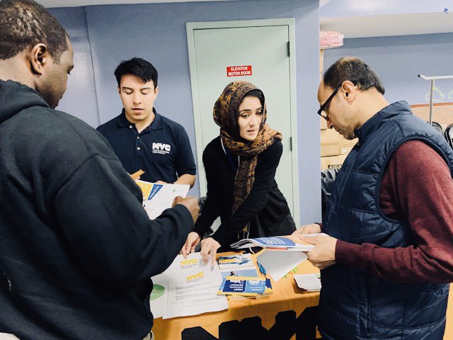 The image shows a man and woman standing behind a table with a golden tablecloth, and two men looking at flyers on the table.