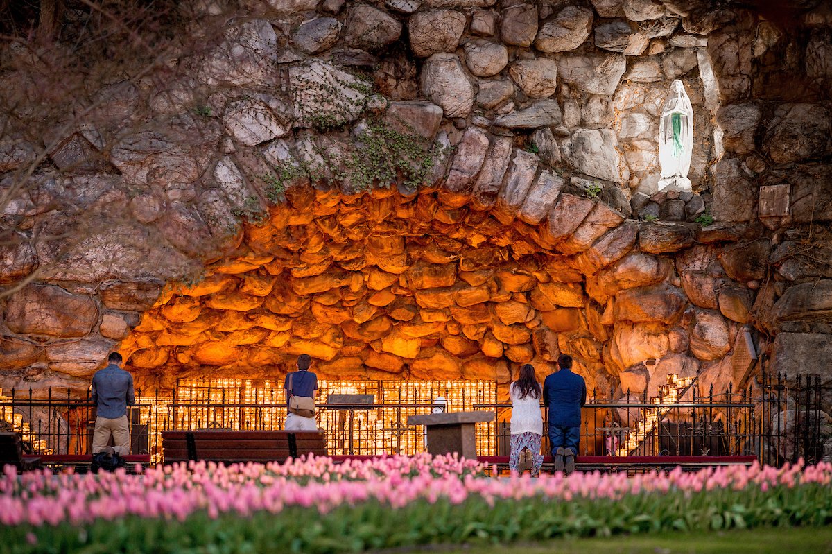Four people kneel in front of candles at the Grotto of Our Lady of Lourdes at the University of Notre Dame