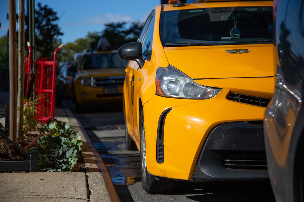 The image shows a yellow taxi on an NYC street.  On the left is a street tree, and a red cart on the sidewalk.  It is a sunny day.