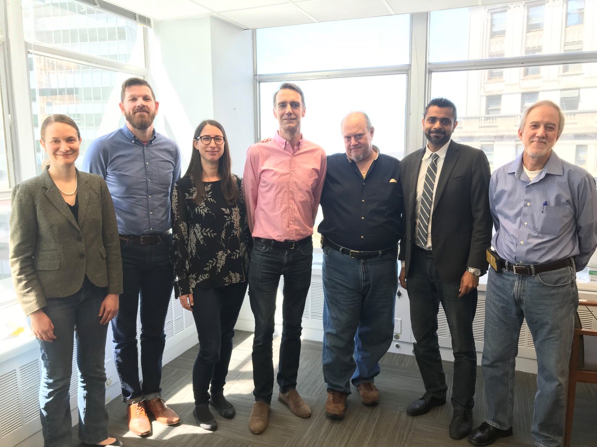 The image shows seven men and women standing together wearing jeans, with large windows behind them.