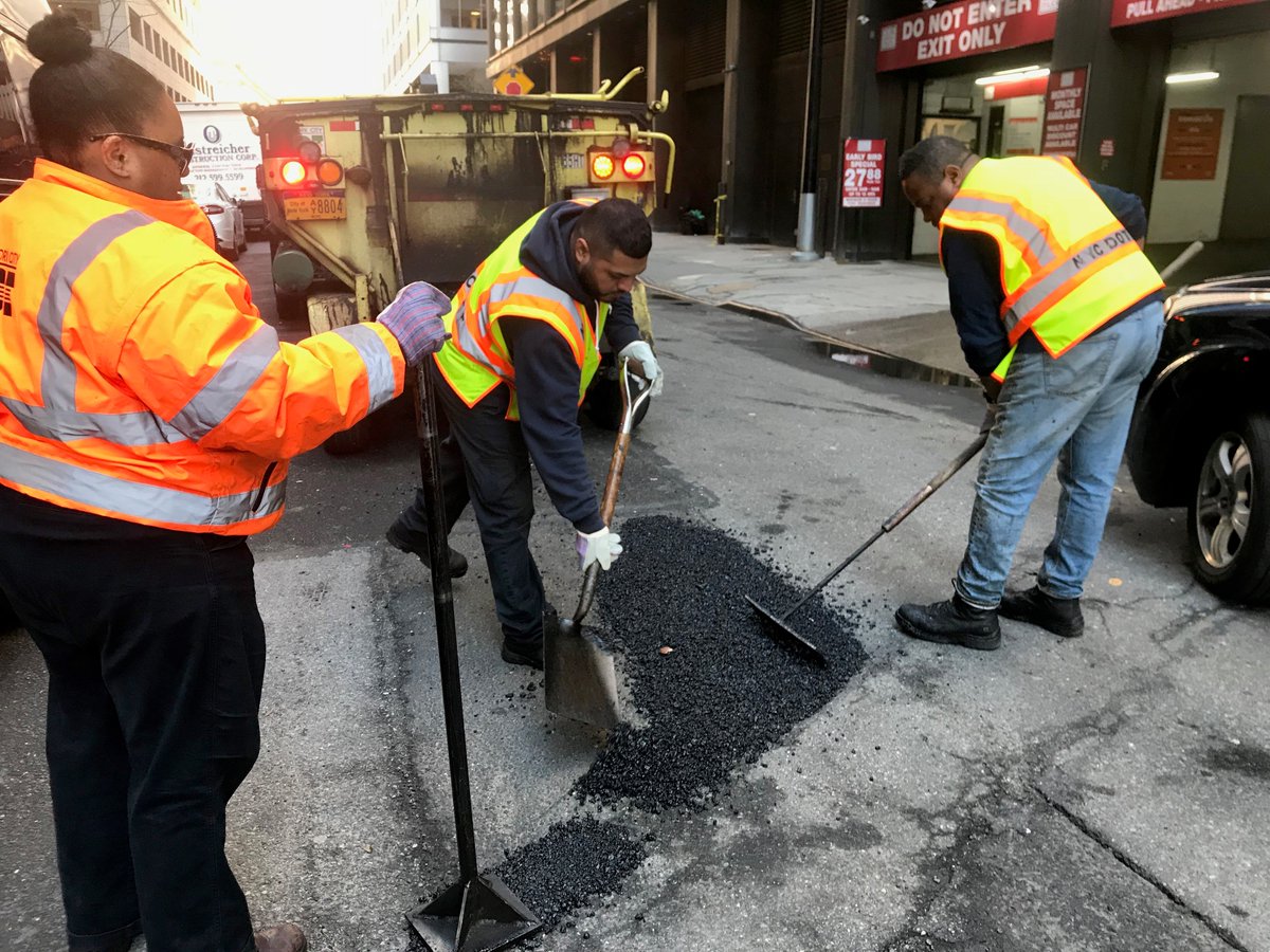 Three people in yellow and orange safety gear fill a pothole with asphalt.