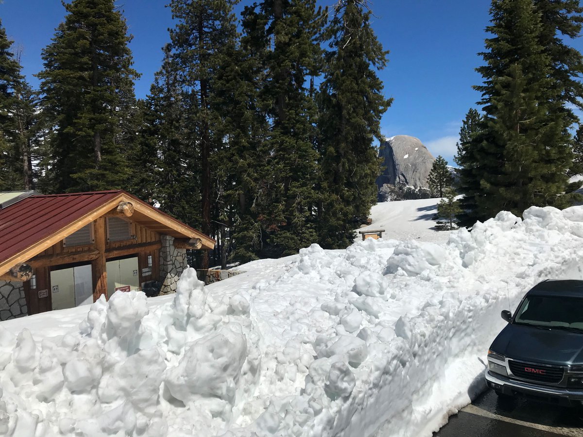 Pickup truck parked next to a wall of snow several feet high. Building protruding from snow. Half Dome in background.