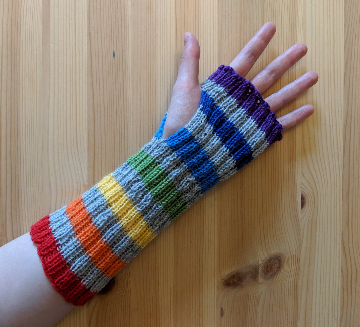 A knitted ribbed armwarmer modelled on a hand/arm, basically a tube with a hole for the thumb. These are longer, with rainbow stripes alternated with light grey stripes. Red, grey, orange, grey, yellow, grey, etc., up to a nice rich purple at the end over the knuckles of the hand.