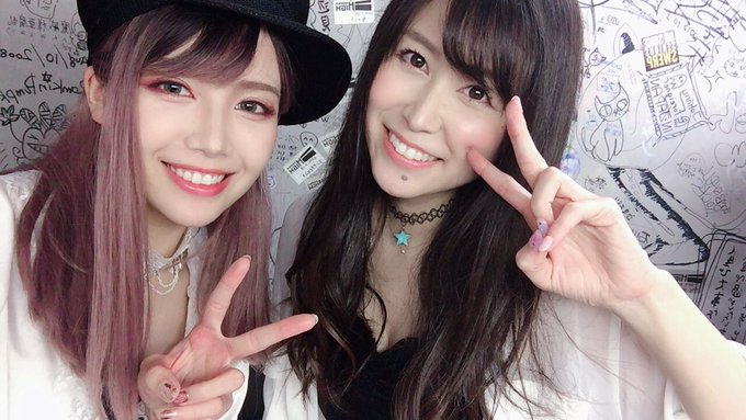 Two young women posing together for a selfie, both making peace signs, against a wall covered in handwritten graffiti and stickers.