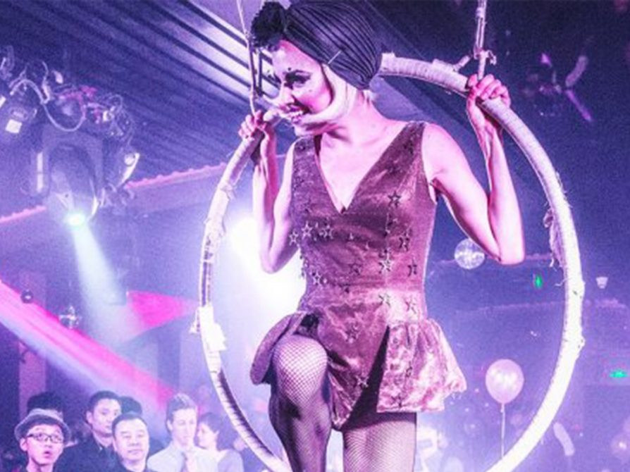 Jones-Rooy within a acrobatic circle suspended over a stage. Her hands are clasped above the shoulder. Her right foot is balanced at the bottom while her left leg points straight down.
