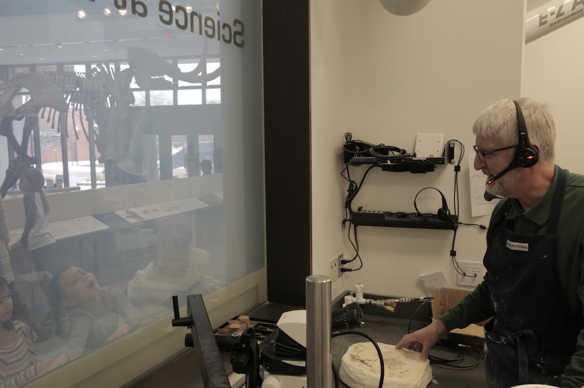 A man wearing a headset in a fossil lab speaks to children on the other side of a large glass window that looks into the lab.