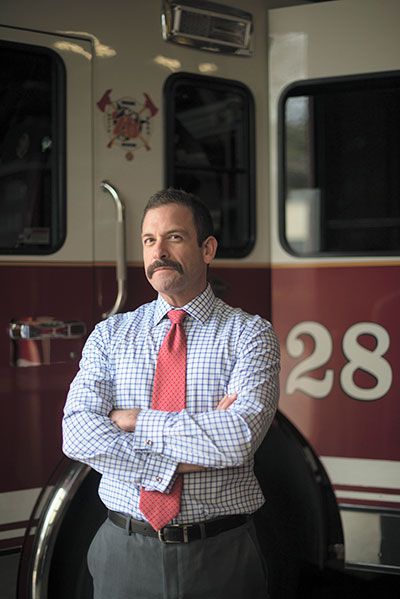 A profile photo of Cristian Hinojosa ’00 standing with his arm's crossed and firetrucks in the background. 