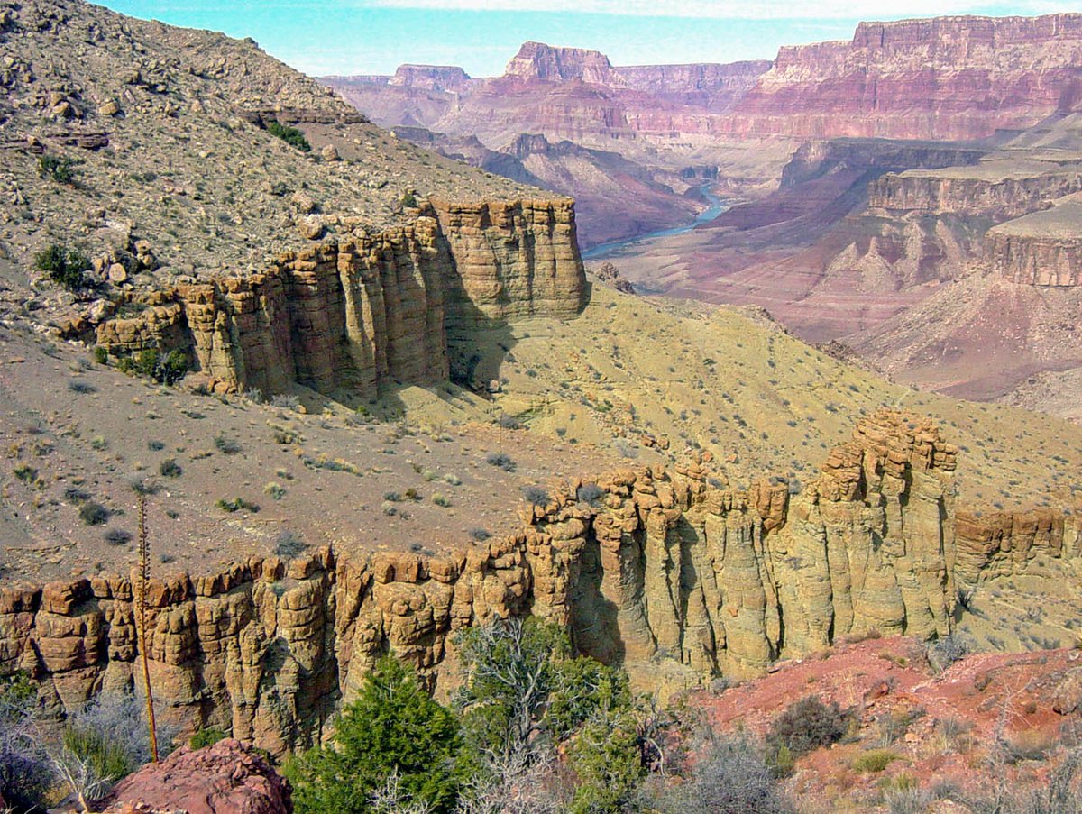 Description: greenish foreground cliffs eroded into irregular columns. In the distance, a river flows between reddish slopes and cliffs.
