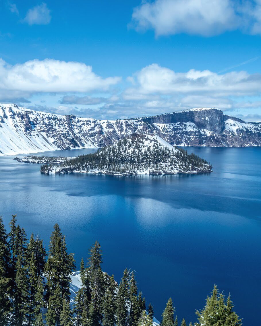 A small island covered in snow and scattered trees sits in a blue lake surrounded by cliffs.