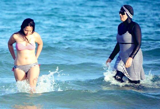 Two women walking in the water at Tunisian beach: one wearing a "burkini", a full-body swimsuit designed for Muslim women; the other wearing a traditional two-piece bikini