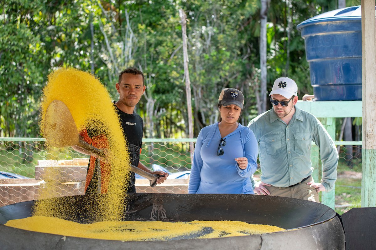 A male and female MBA student examine farinha, a starch product made from mandioca root, while a man throws some into the air while stirring the starch in a large pot.