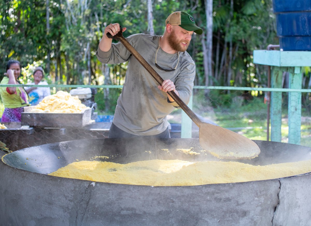 An MBA student wearing a Notre Dame baseball cap stirs a steaming vat of farinha