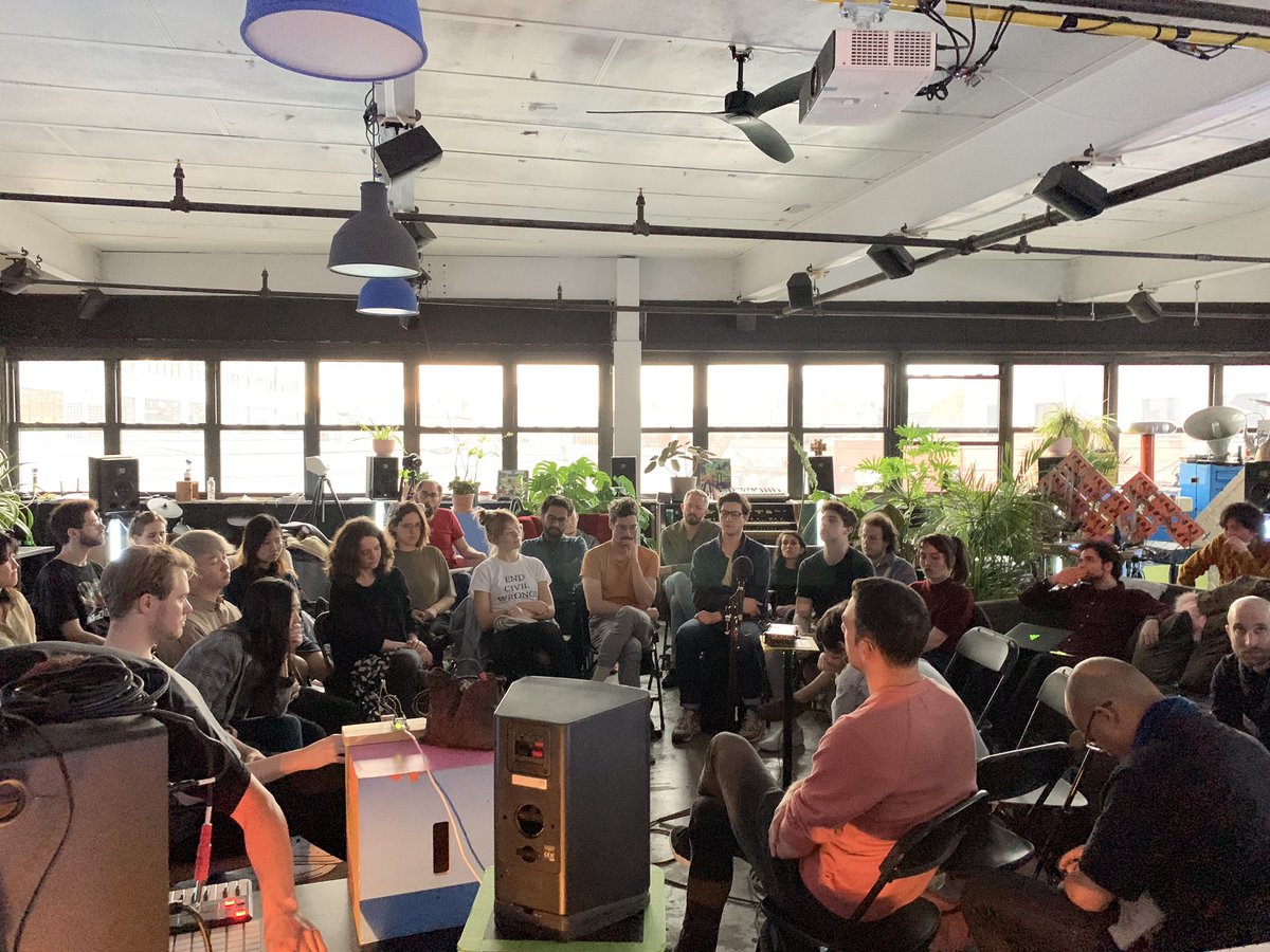 a couple dozen people sitting in a ring surrounded by speakers at ear level and in the ceiling