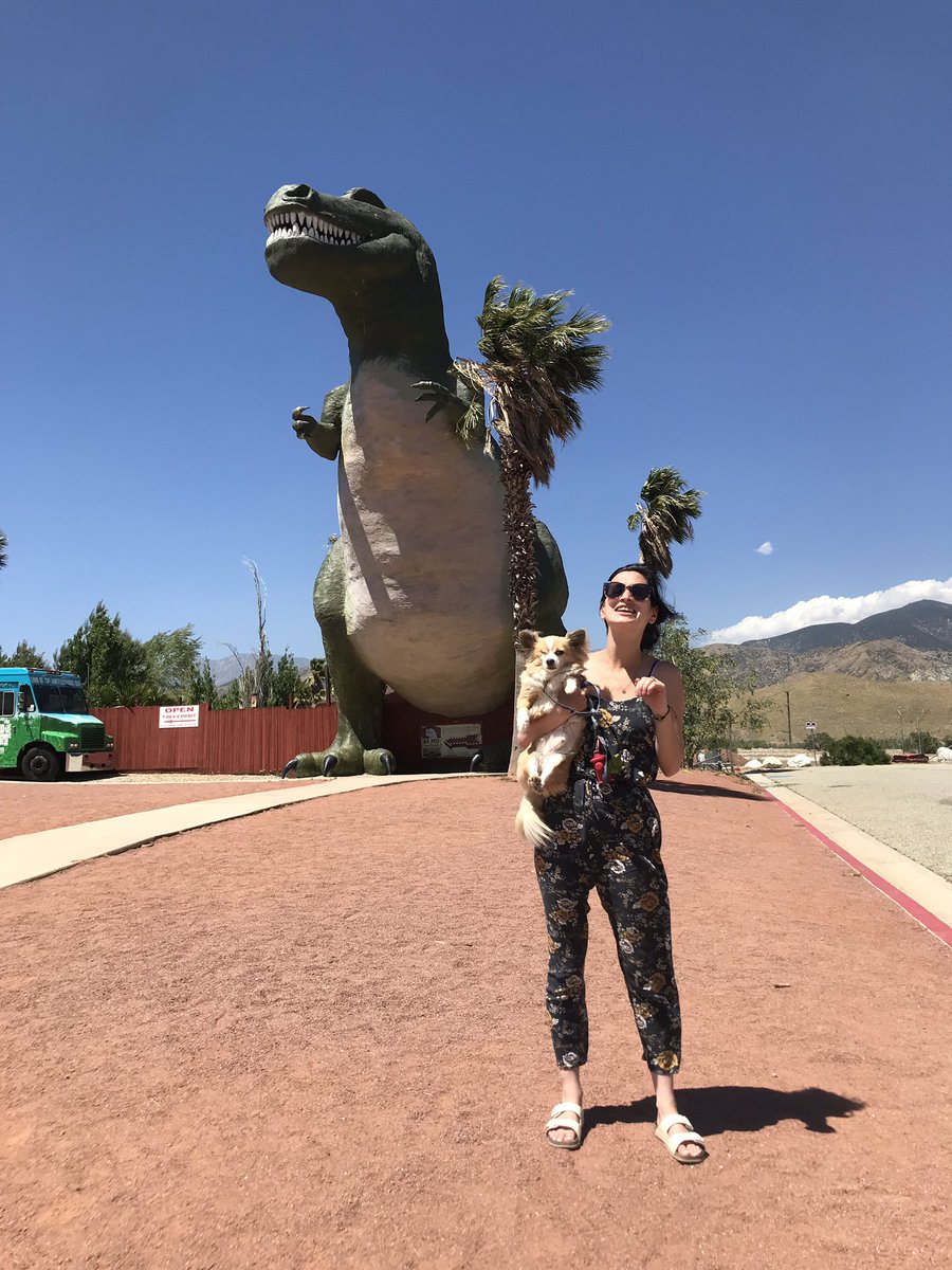 Bea (brunette white woman) standing in front of a giant T. rex statue holding Fitz the one eyed dog trying to get him to hold his little paws up like the dinosaur