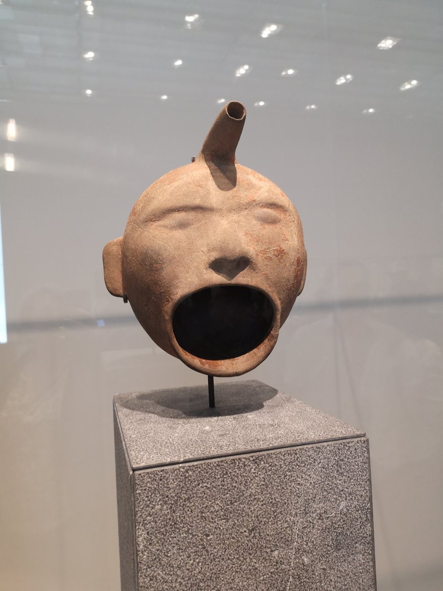 A vase in the shape of a head. There is a spout from the top and the mouth of the face is a wide oval.