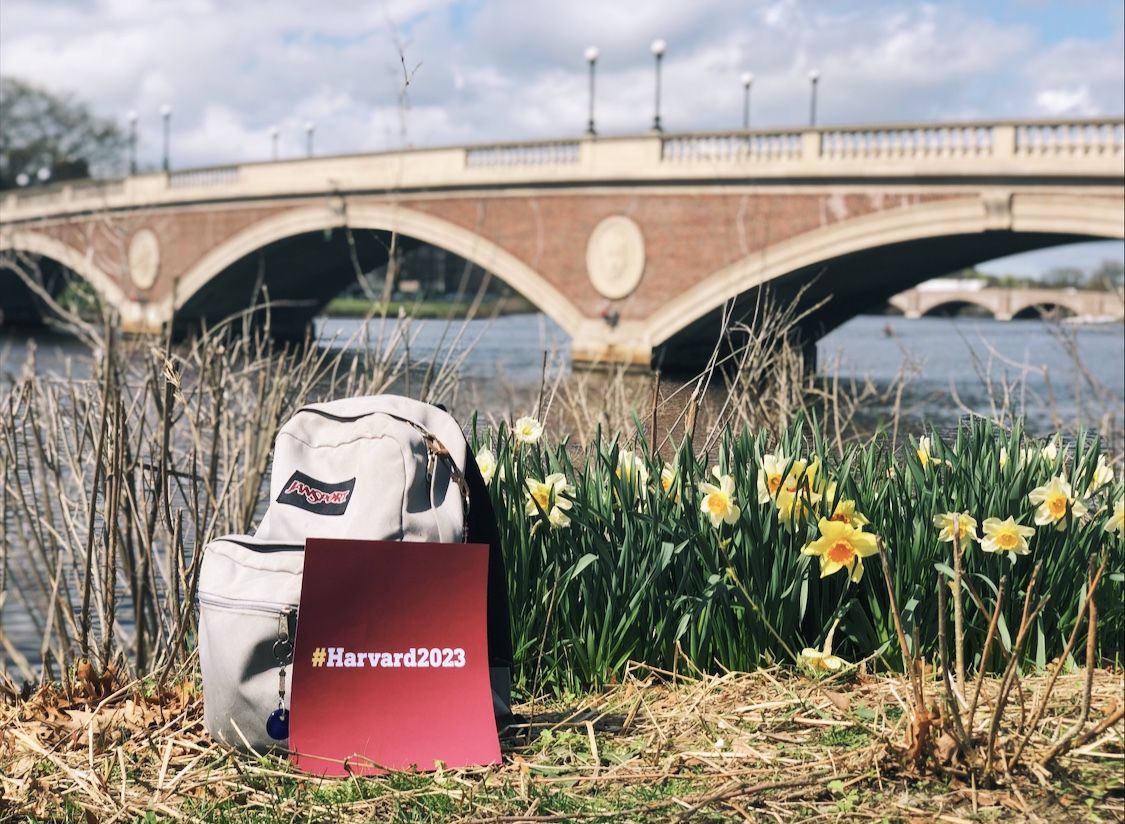 A backpack, flowers, and a sign that says "#Harvard2023" by the Weeks Bridge 