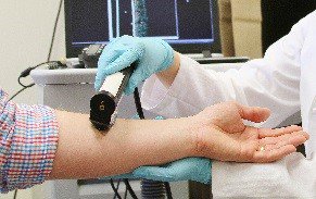 Image: a dermatologist examining a patient’s arm with an instrument.