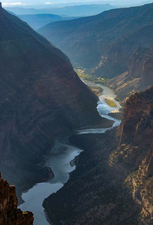 A view down into a steep rock canyon with a narrow river curving along the bottom far below moving from sunlight to shadow.