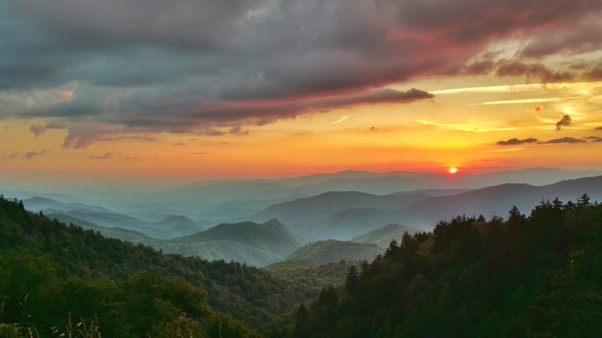 Layers of rolling green mountains stretch out to the horizon under an orange sunset sky.