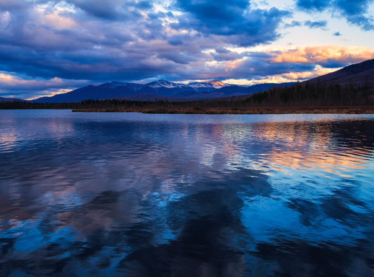 A calm, flat pond is bordered by rising hills with a line of mountains on the horizon under a stormy sky.