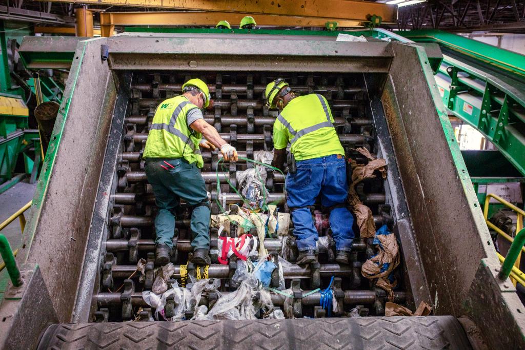 Two workers remove plastic bags that have jammed machinery at a recycling center