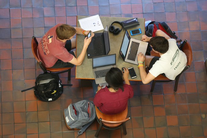 Three students study together at a table, with laptops, phones and backpacks.