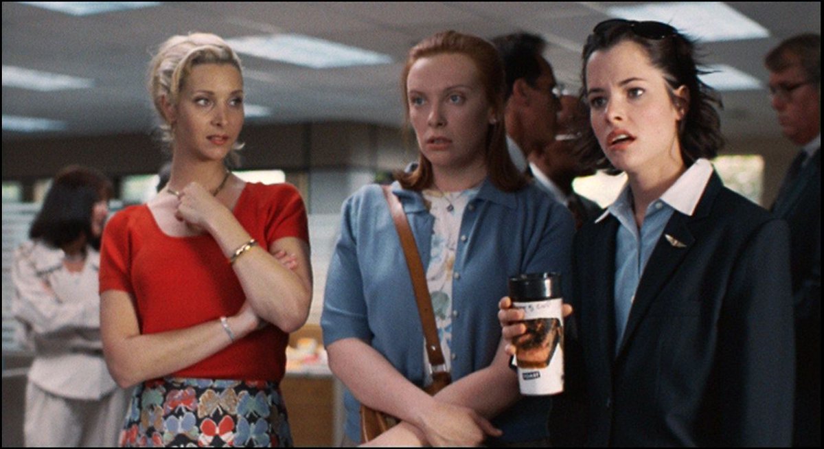 Image from the film 'Clockwatchers' (1997). Three women stand in an office building, reacting to something out of frame. The first woman looks to the other two, uncertain, while the other women look to be in two varying states of shock.