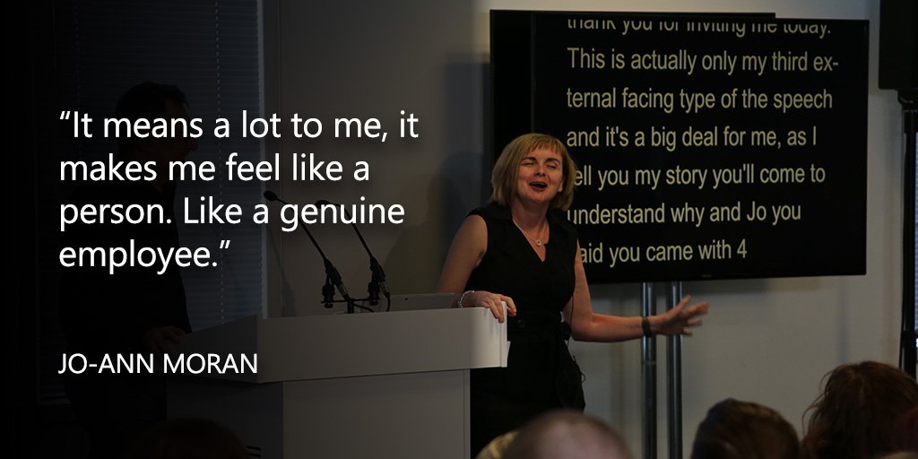 Photography of a woman wearing a black dress. She is talking to a live audience with her hand resting on a white plinth. In the background there is a large television screen showing subtitles.