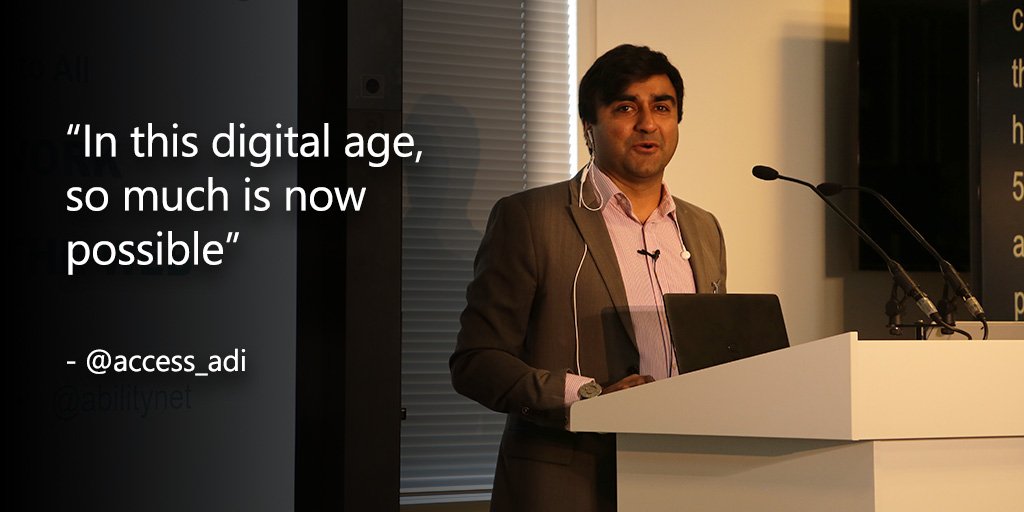 A photograph of Adi, the first guest speaker from Ability Net. Adi is wearing a smart jacket and shirt, speaking into microphones on a white plinth. 