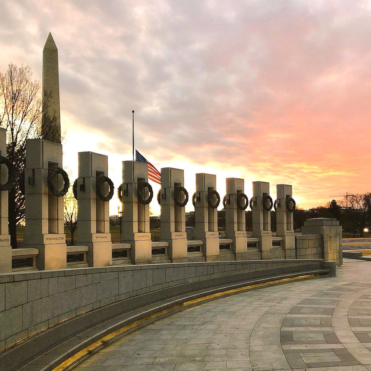 A line of stone pillars stand along an open plaza with the tall tower of the Washington Monument in the background.