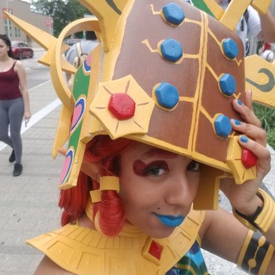 A closeup of a cosplayer dressed as Riju from Legend of Zelda: Breath of the Wild. They are holding a prop version of the Thunder Helm over their head and are peeking out from under it to smile at the camera.