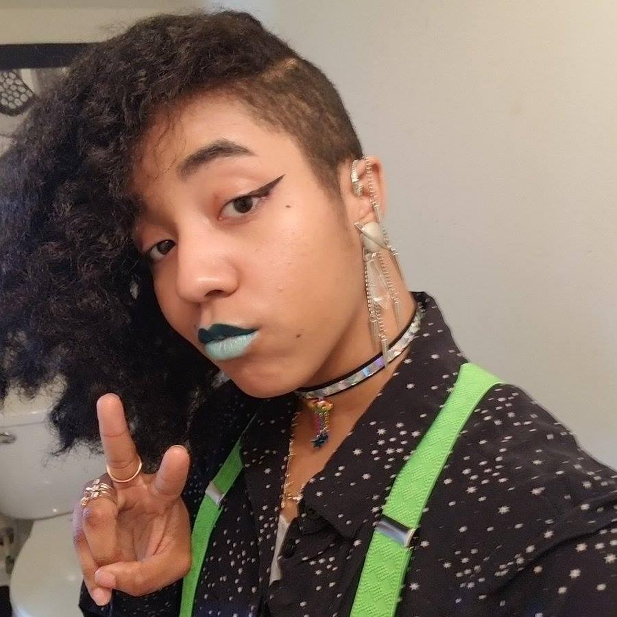 A black non-binary person with an undercut. The rest of their hair is swept to the side. They're wearing numerous accessories, dual tone lipstick, and bright clothing. They are looking at the camera with a V pose.
