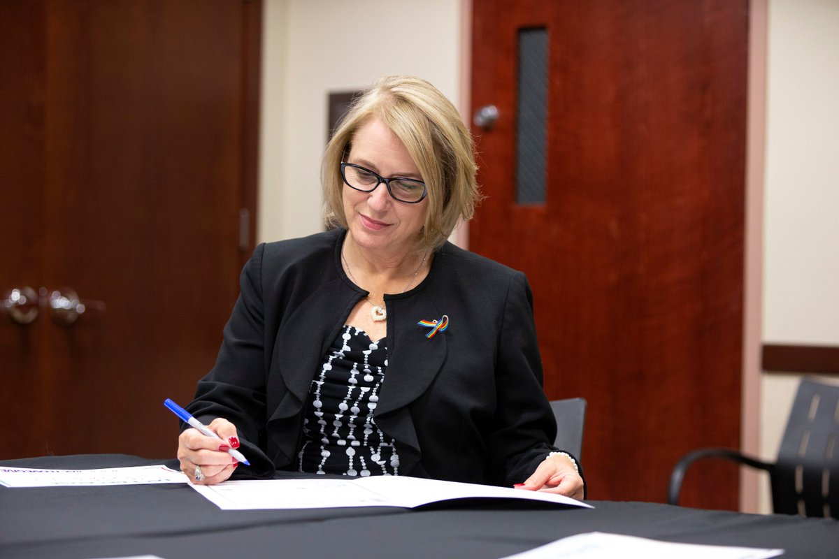 Commissioner VanderLey signing a proclamation, inside, sitting at a table with a black tablecloth. 