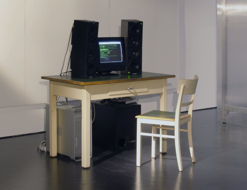 Atop a spare blonde desk sits a black CRT monitor and large speakers, below further speakers and a steel desktop computer, nearby a chair