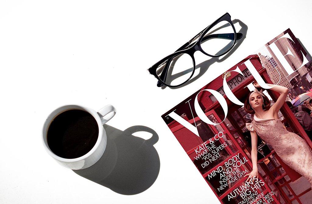 an overhead shot of a vogue magazine, coffee cup and a pair of glasses on a taple top