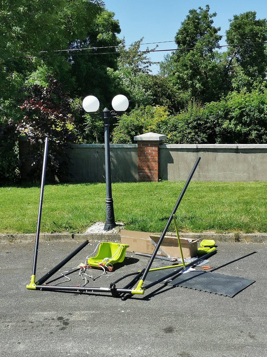 Picture of a swing set being put together in a garden. Blue skies surrounded by large trees. A patch of green grass behind the swing and a wall.