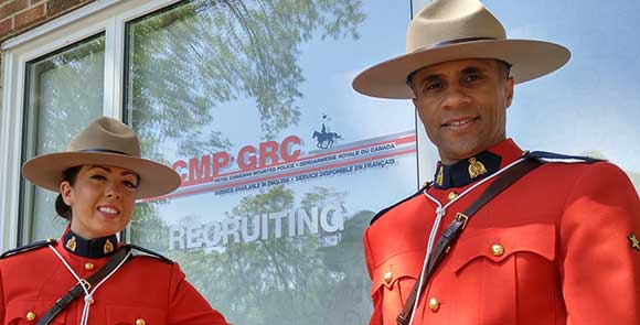 Une policière et un policier de la GRC en tunique rouge, debout devant un immeuble, avec, en arrière-plan, un panneau de recrutement bilingue de la GRC. 