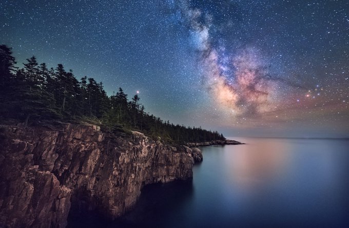 The long band of stars making up the Milky Way hangs in the night sky above the ocean and a rocky coastline.