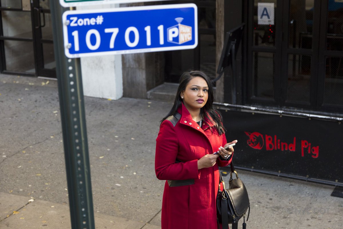a blue zone sign with numbers is posted to a sign post on the sidewalk. a woman in a red jacket looks up at the sign while holding her car keys and cell phone.