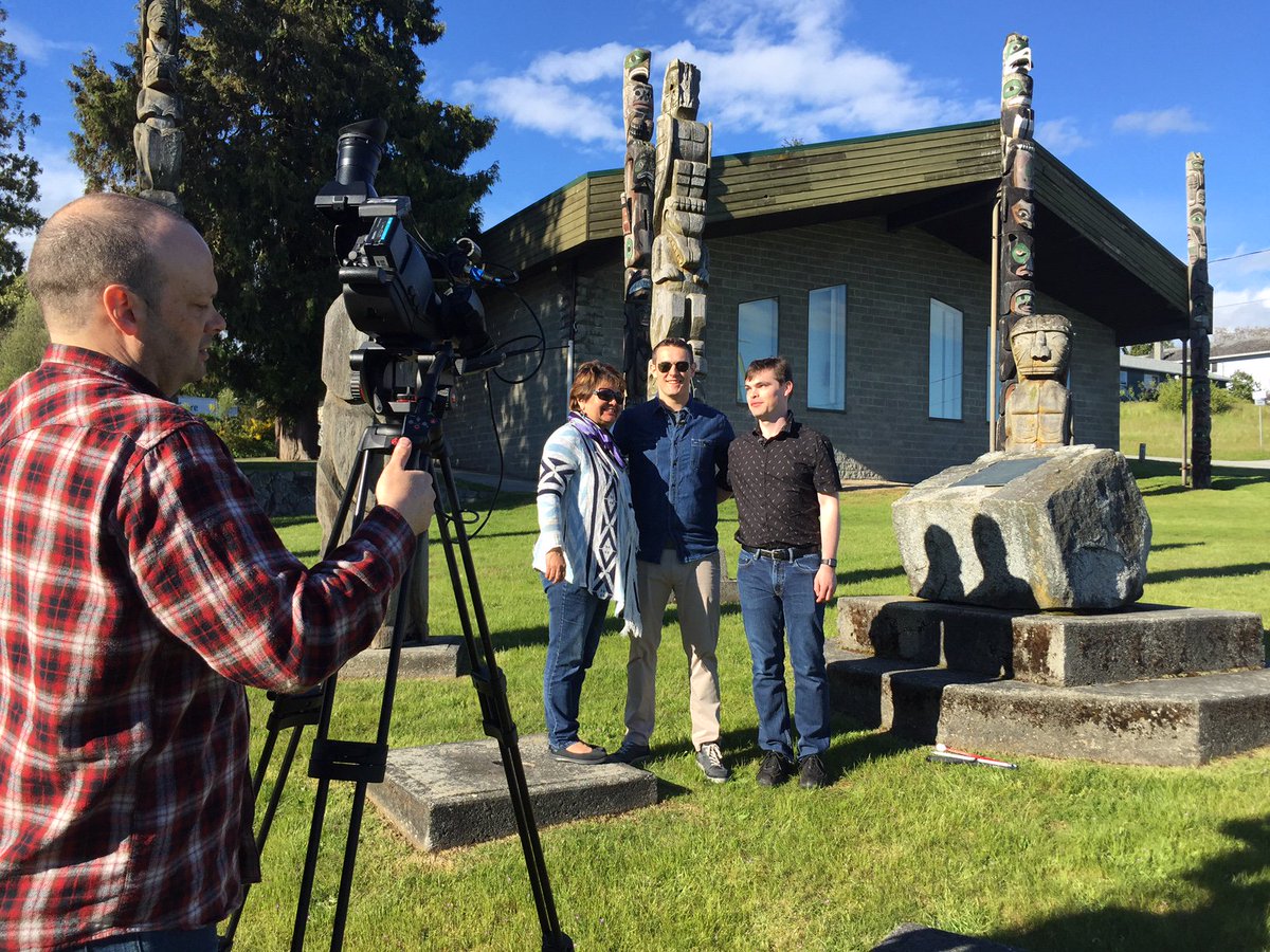 A camera films Anthony McLachlan, Grant Hardy and a female guest. They are surrounded by traditional totem poles on a very sunny day.