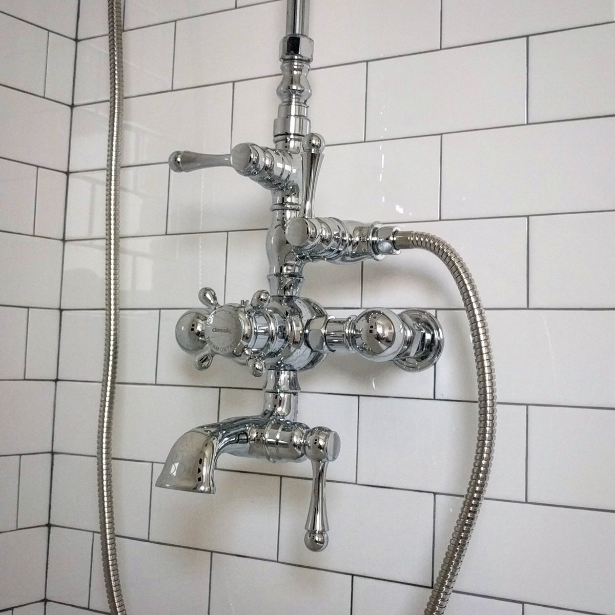A complex, steam-punk looking shower faucet. It has one dial and three levers, each coming out at on a different axis. It is all mounted outside of the wall, set away from it by a couple inches. There is also a hose coming out out of it and heading out of the frame. The faucet contraption is chrome, the wall behind it is white subway tile.
