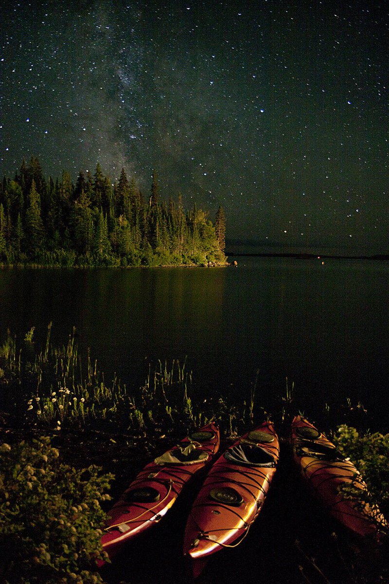 Three red kayaks rest on the bank of a lake at night with a tree-covered island in the background and the Milky Way overhead
