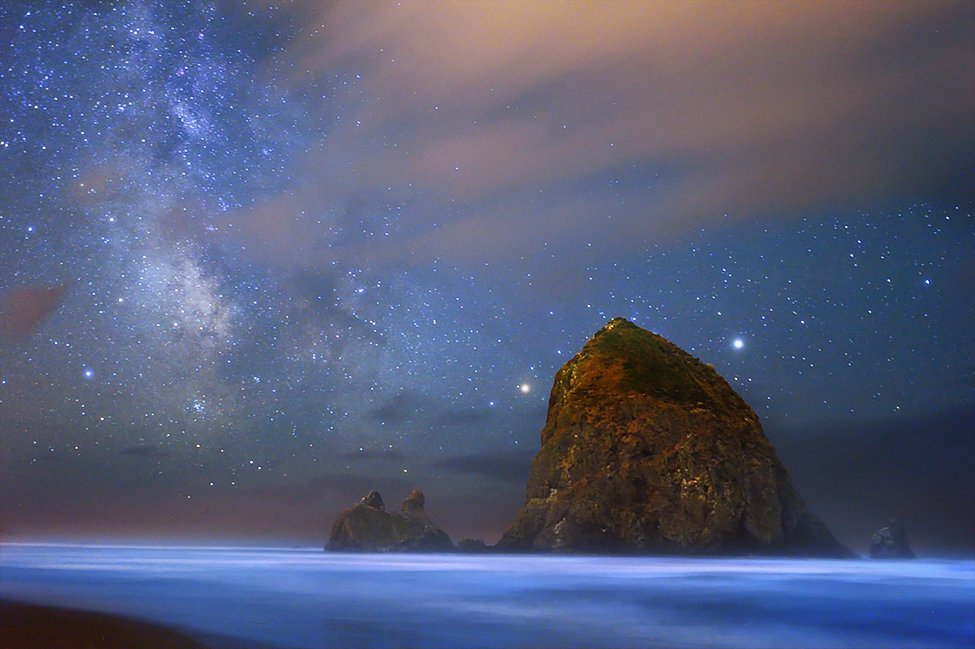 With a large rock just offshore, shallow water along a sandy beach glows with blue light under a starry sky.