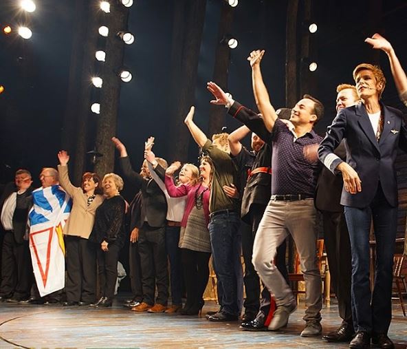 Photo of the cast of We Come From Away waving to spectators. A castmember on the far left is holding up the Newfoundland flag.