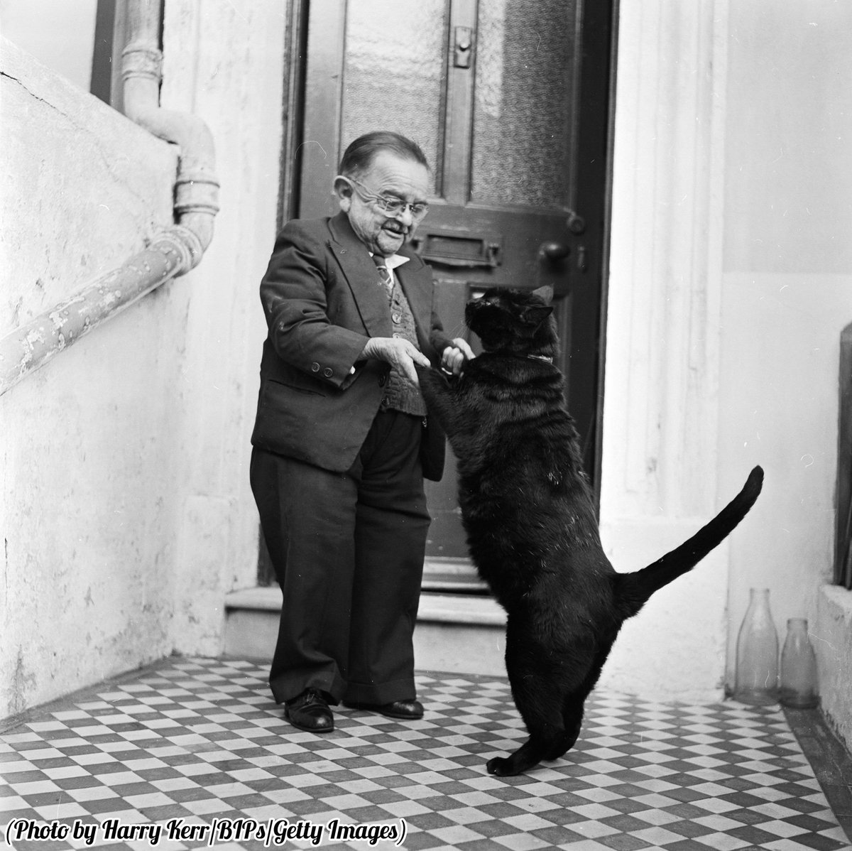 Henry Behrens, the smallest man in the world dances with his pet cat in the doorway of his Worthing home, 1956.