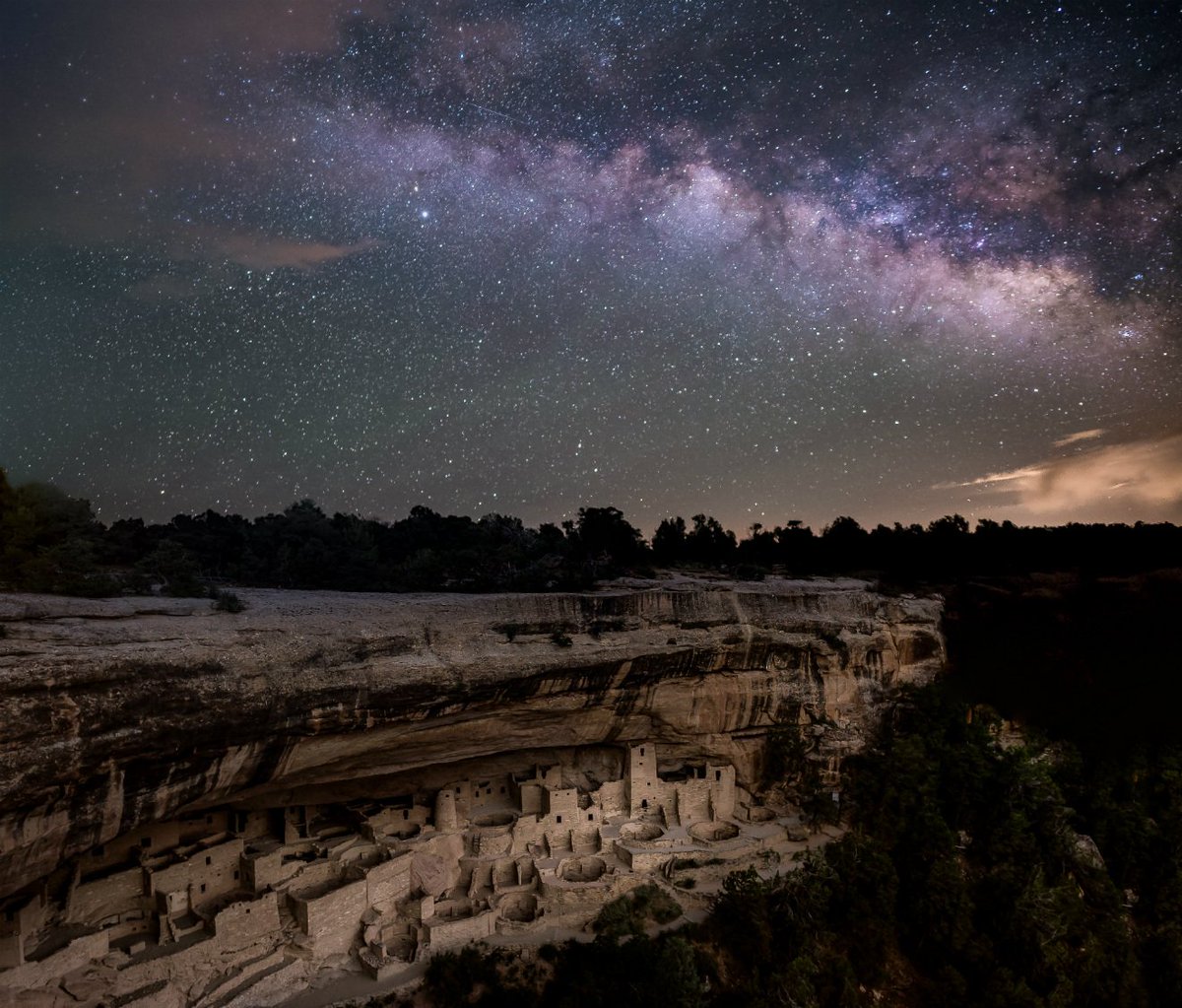 The ancient remains of a brick walled city cling to the wall of a cliff under a rock overhang while the stars of the Milky Way shine from above.